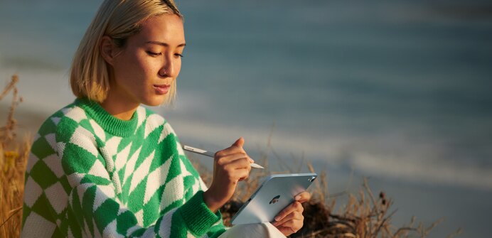 A person on the beach holding their Apple Pencil Pro in one hand and a blue iPad mini in the other